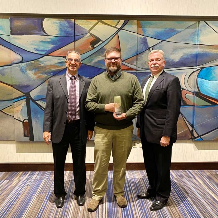 Standing from left to right in front of an abstract art piece: Ray Phillips, Chairman of the Board; 2019 Cushman Award winner Frank Ferdinand, Service Desk Agent; and David Sprague, President/CEO.